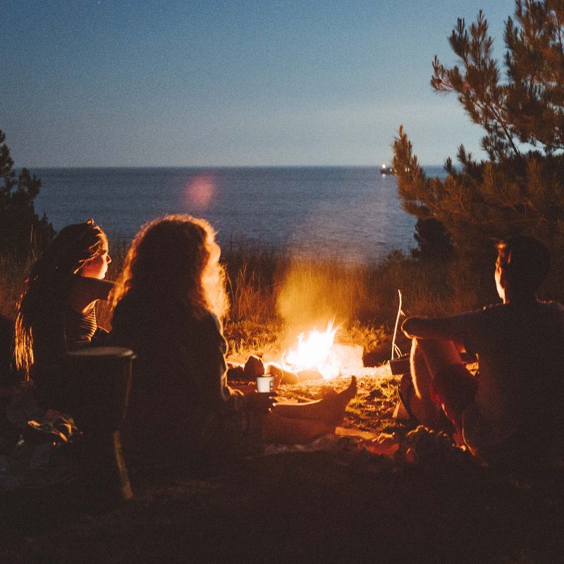 an outside fire surrounding by people with a lake in the background